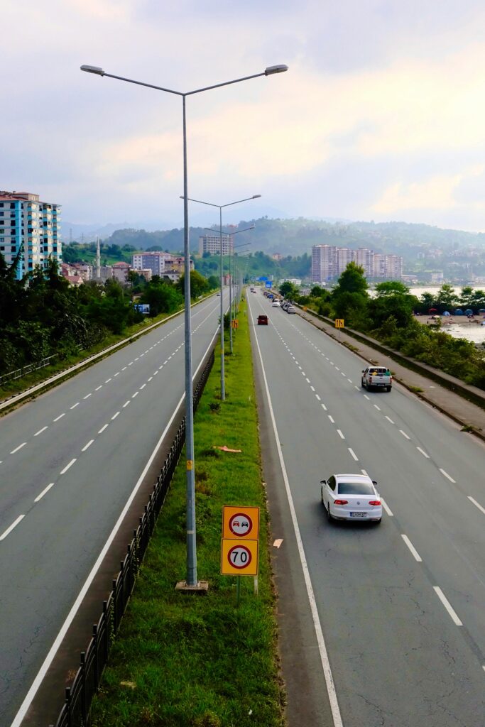 pexels-photo-33730778-33730778 Aerial view of a calm highway with sparse traffic amid lush greenery and distant buildings.
